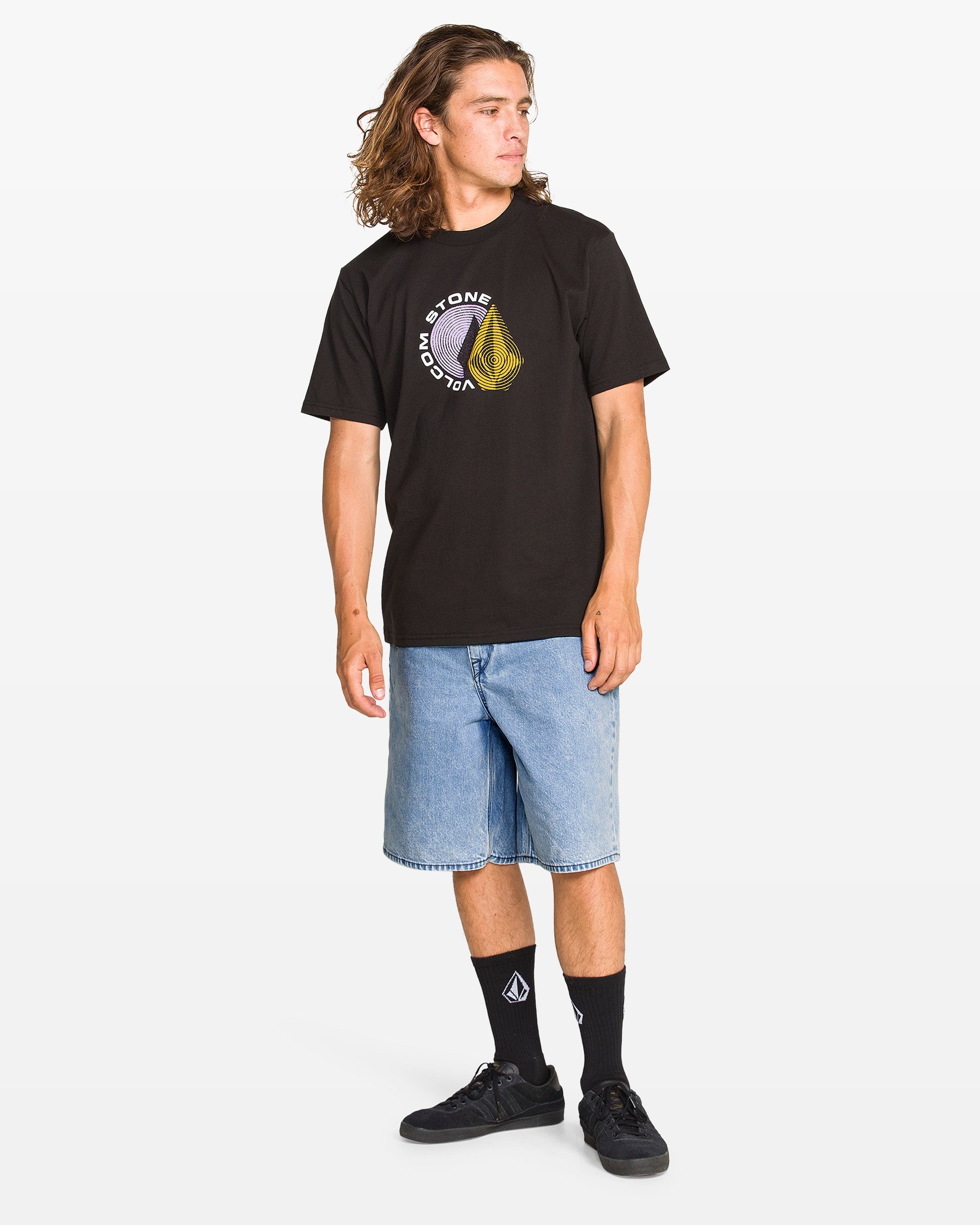 A person with long hair wears a black graphic tee, Volcom Men’s Billow Denim Shorts in light blue recycled cotton, black logo socks, and black sneakers, standing against a plain white background.