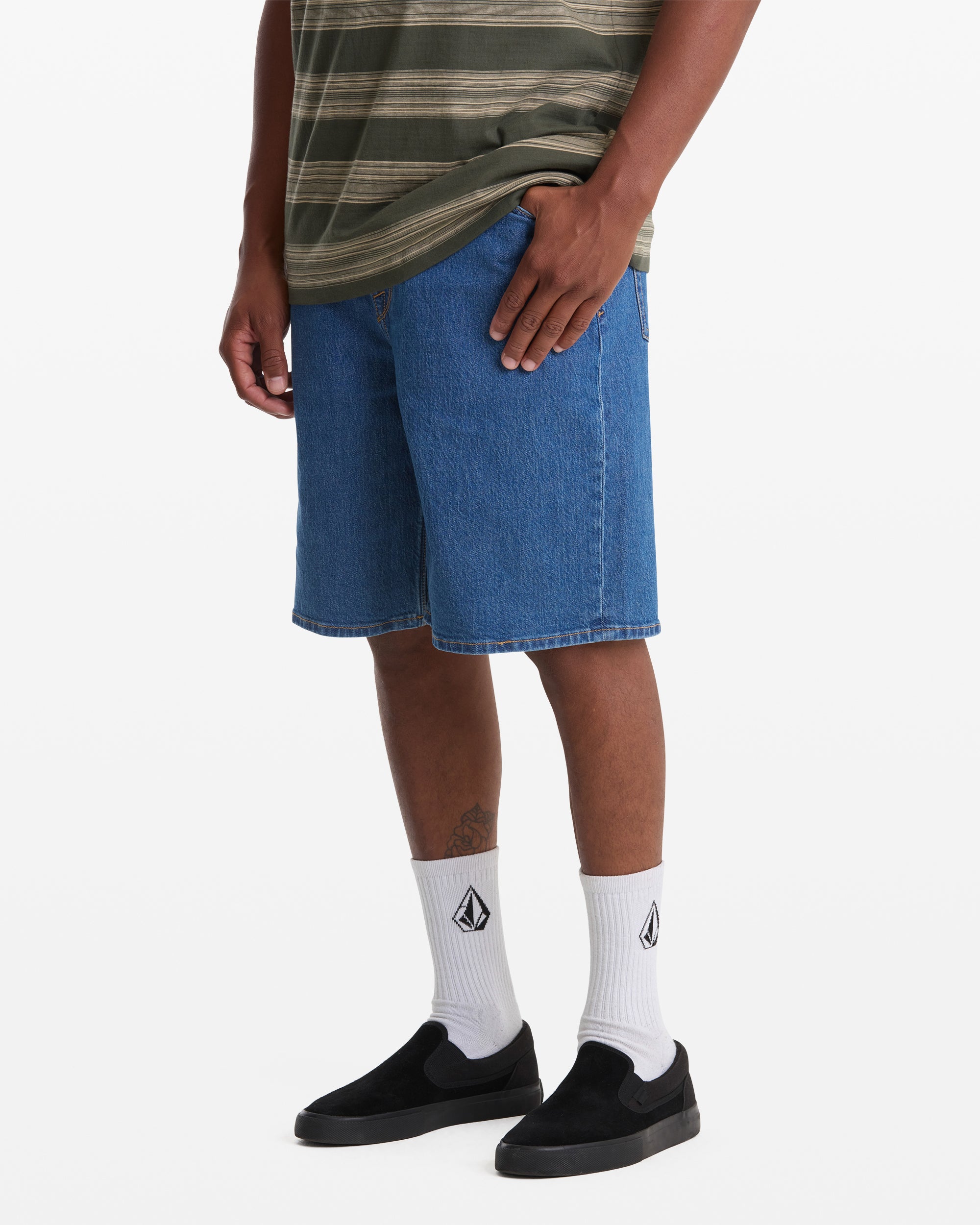 A person is shown from the waist down wearing Volcom's Men's Billow Denim Short 22, a green striped t-shirt, white socks with a black logo, and black slip-on shoes, standing against a white background.