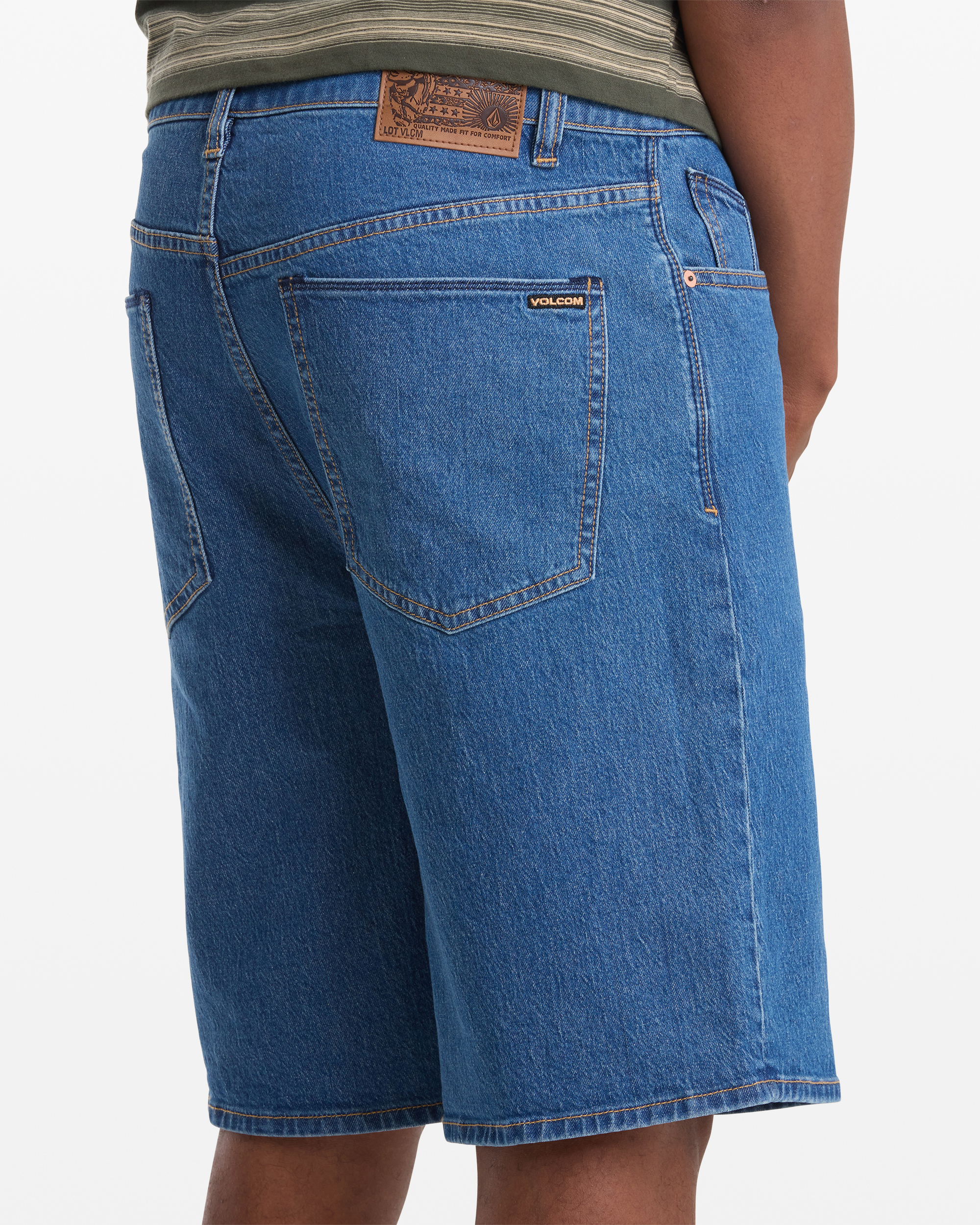 A person is seen from the back wearing Volcom Men's Billow Denim Short 22, featuring blue denim, large rear pockets, a leather waistband patch, and visible stitching—paired with a green striped shirt.