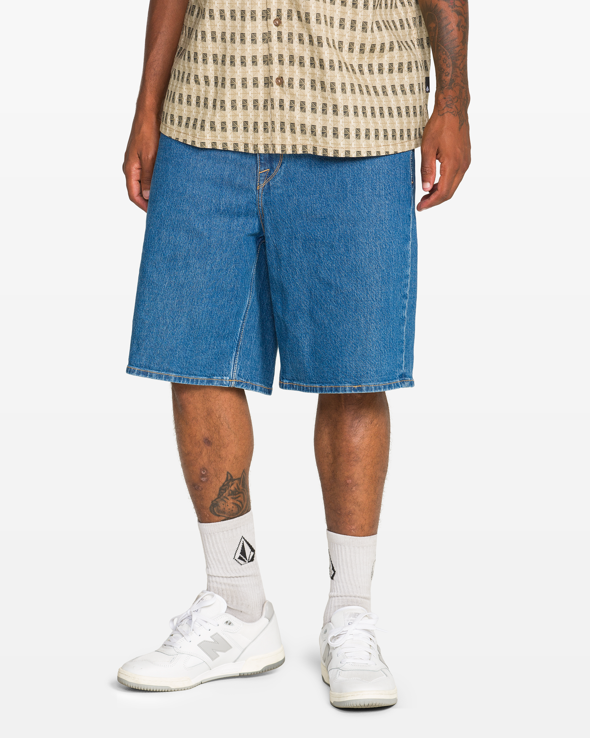 Only the lower half of a person is visible, wearing Volcom Men's Billow Denim Short 22 in blue, a beige patterned shirt, white crew socks with a black logo, and white sneakers.