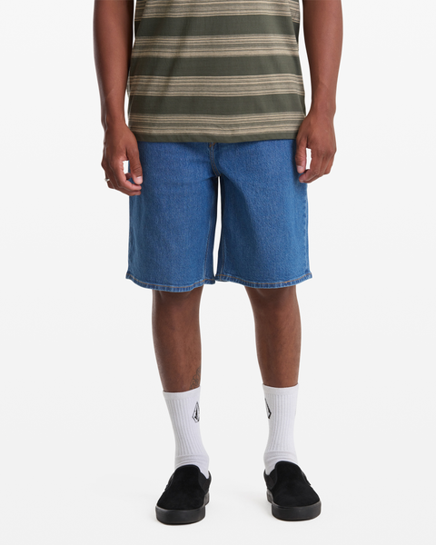 A person wearing a green striped shirt, Volcom Men's Billow Denim Short 22, white socks with a logo, and black slip-on shoes stands against a plain white background.
