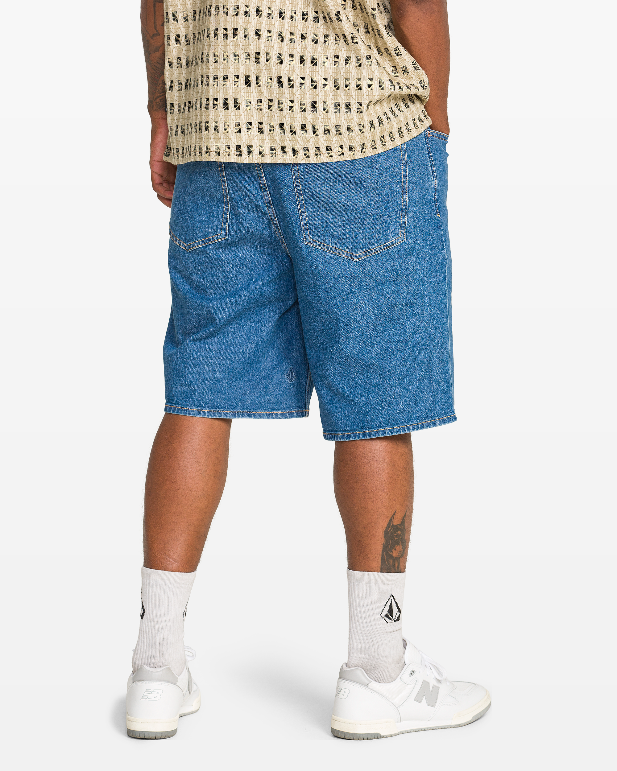 A person stands facing away, wearing a beige patterned shirt, Volcom Men's Billow Denim Short 22 in blue, white socks with a black logo, and white sneakers. A tattoo is visible on the back of their left calf.