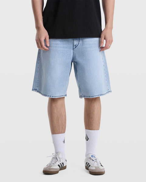 A person stands against a white background wearing Volcom Men's Billow Denim Shorts in light blue, a black oversized-fit shirt, white crew socks with black logos, and white Adidas sneakers with black stripes.