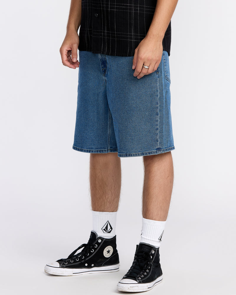 A person stands wearing knee-length Volcom Men's Billow Denim Shorts, a black plaid shirt, white logo crew socks, and black high-top Converse sneakers. The background is plain white, and the person’s face is not visible.