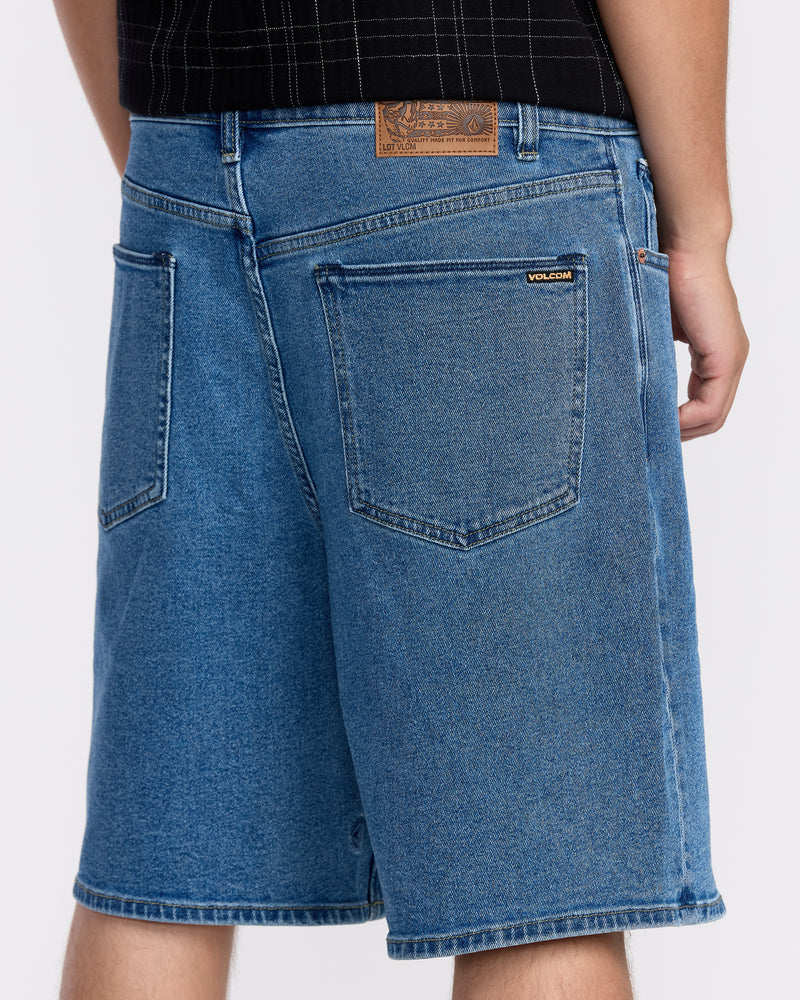 A person wears Men's Billow Denim Shorts by Volcom, featuring visible back pockets and a brown leather patch on the waistband, made with Water Aware construction, standing against a plain background.