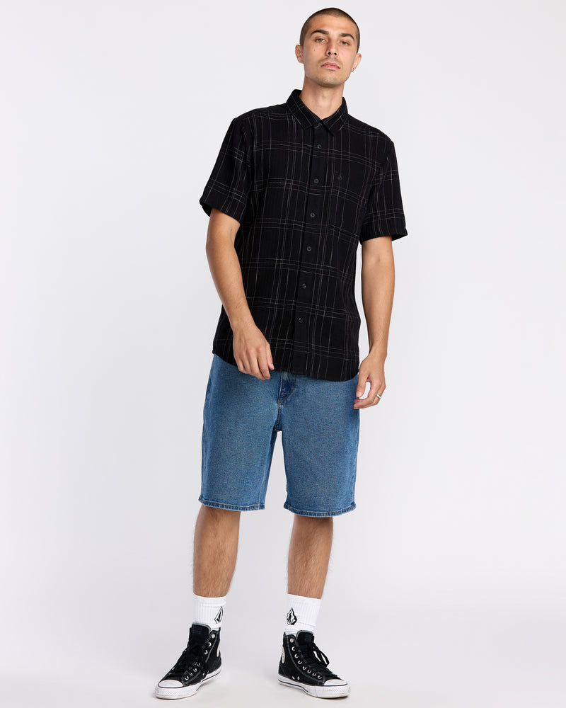 A man stands against a plain background wearing a black plaid short-sleeve button-up shirt, Volcom Men's Billow Denim Shorts with Water Aware construction, white socks, and black high-top sneakers.