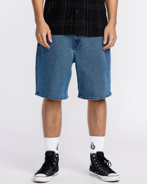 A person wearing blue Volcom Men's Billow Denim Shorts with Water Aware construction, a black plaid short-sleeve shirt, white socks with a black logo, and black high-top sneakers stands against a plain white background.