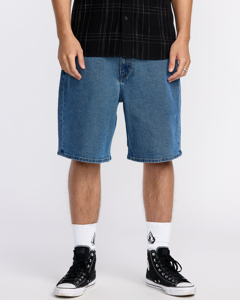 A person wearing blue Volcom Men's Billow Denim Shorts with Water Aware construction, a black plaid short-sleeve shirt, white socks with a black logo, and black high-top sneakers stands against a plain white background.