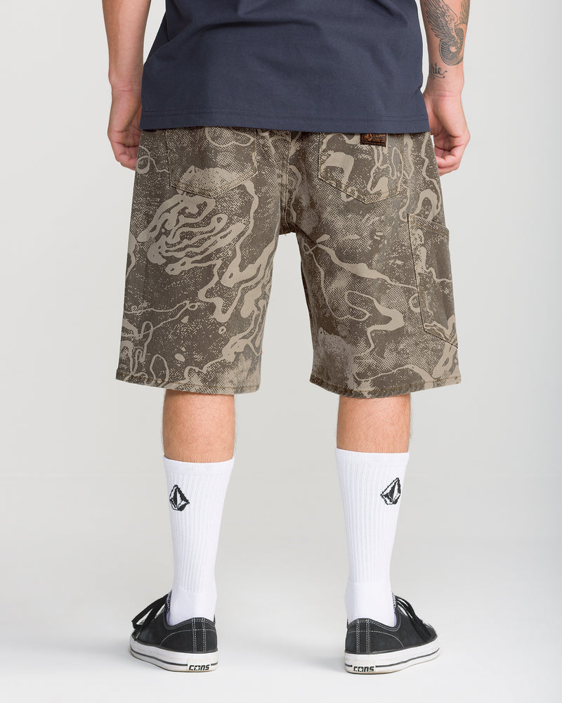 Back view of a person wearing Volcom Men’s Kinkade Utility Denim Short 22 in knee-length khaki camo with an abstract brown pattern, white crew socks, and black sneakers.