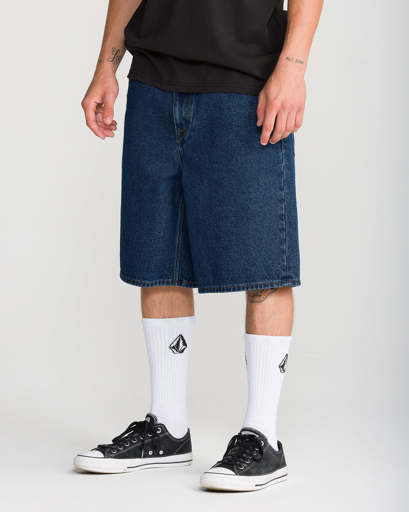 A person wearing dark indigo Volcom Men’s Billow Denim Short 22, white crew socks, and black low-top sneakers.