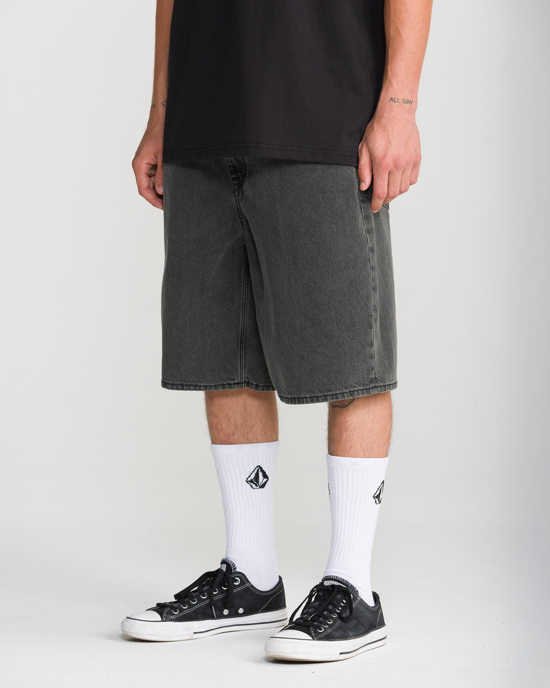Volcom Men’s Billow Denim Short 22 in black, featuring a loose fit falling to the knees, worn with white crew socks and black low-top sneakers.