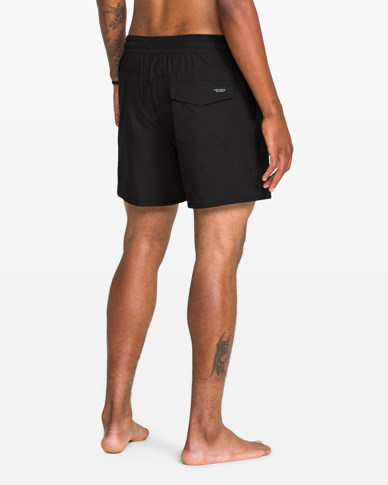 A person stands barefoot, facing away, wearing Volcom Men's Lido 2 Solid Trunks made from recycled polyester with a back pocket. Visible tattoos are on their right arm and left leg, against a plain white background.