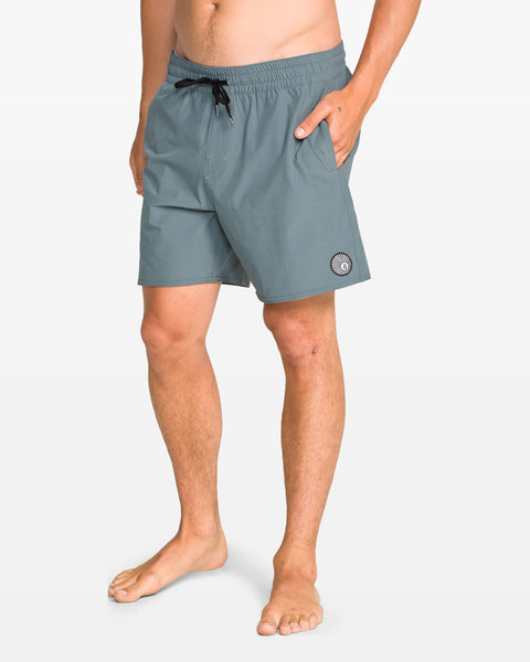 A man stands barefoot in Volcom Men's Lido Solid 16" Trunks—light teal boardshorts with a 5-inch inseam, drawstring waist, pockets, and a circular patch—made from recycled polyester and set against a plain white background.
