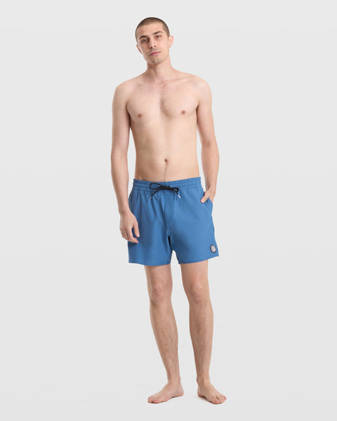 A man with short hair stands barefoot against a white background, wearing Volcom Men's Lido Solid 16" Trunks made from recycled polyester, featuring a small round logo on the left leg.