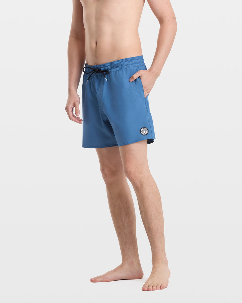 A man stands barefoot against a white background, wearing Volcom Men's Lido Solid 16" Trunks in blue with a round logo patch by the left hem. His upper body is bare, and one hand is in his pocket.