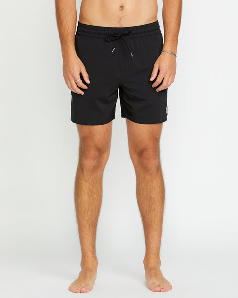 A barefoot model stands against a plain white background, wearing Volcom Men's Lido 2 Solid Trunks in black, made from recycled polyester. The image is cropped below the chest, with the face not visible.