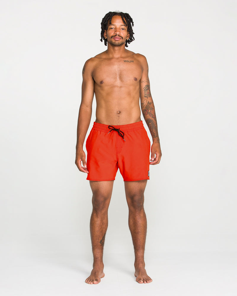 A man with short dreadlocks stands facing forward, shirtless and displaying tattoos on his chest and left arm, against a plain white background. He is wearing Volcom Men's Lido Solid Trunks in a vibrant red color with a 16-inch outseam. These trunks are crafted from recycled polyester, combining sustainability with style.