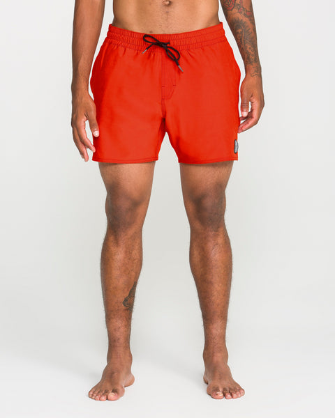 A person stands barefoot wearing bright red Volcom Men's Lido Solid Trunks, made from recycled polyester with a black drawstring and a contemporary 16-inch outseam, photographed against a plain white background.
