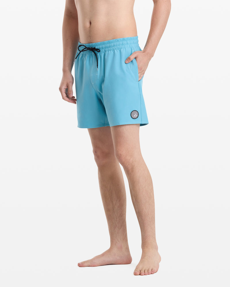 A man stands barefoot against a white background, wearing Volcom Men's Lido Solid Trunk 16 swim trunks in light blue with a black drawstring and a small circular patch on the left leg.