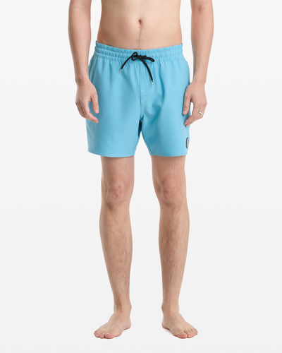 A man stands barefoot against a white background, wearing Volcom Men's Lido Solid Trunk 16 swim trunks in light blue with a black drawstring and small round logo on the left leg. His upper body is not visible.