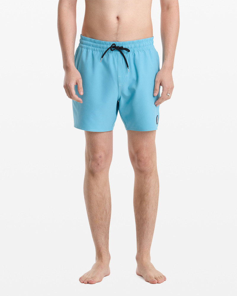 A man stands barefoot against a white background, wearing Volcom Men's Lido Solid Trunk 16 swim trunks in light blue with a black drawstring and small round logo on the left leg. His upper body is not visible.