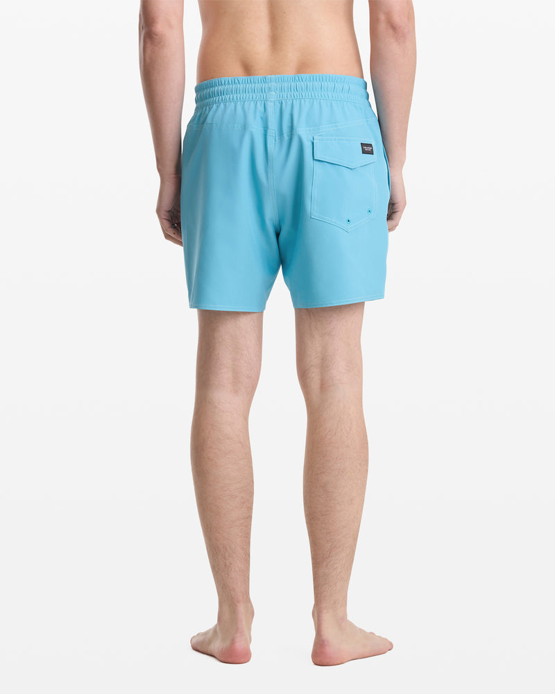 A barefoot person, seen from behind, wears Volcom Men's Lido Solid Trunk 16 swim shorts in light blue with an elastic waistband and a back pocket. The background is plain white.