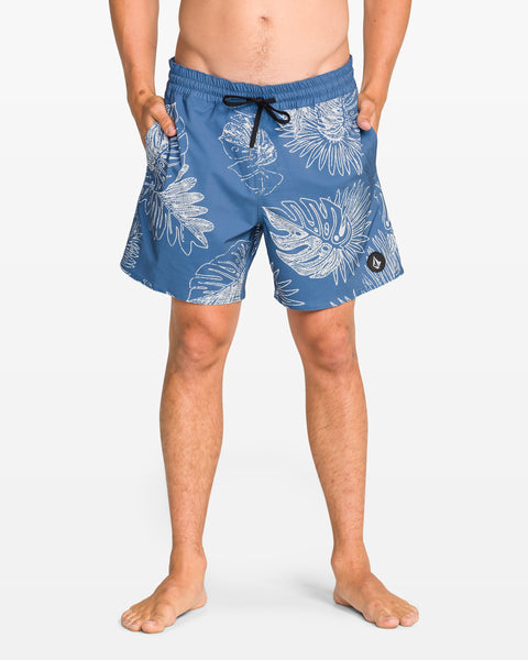 A person stands barefoot with hands in pockets, wearing Volcom Men's Lido Print 16" Trunk swim shorts that feature a blue and white tropical leaf pattern, black drawstring, and 4-way stretch, set against a plain white background.