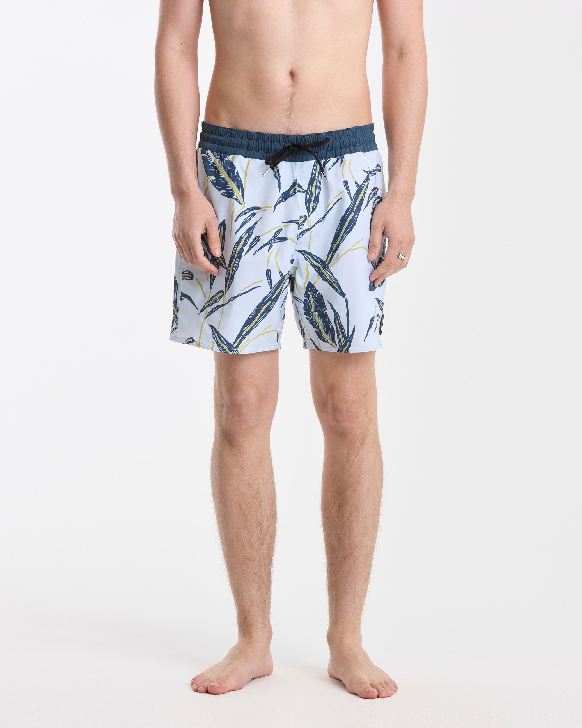 A person stands barefoot against a white background, wearing Volcom Men's Lido Print 16" Trunks featuring a dark blue waistband, leafy blue, green, and yellow print, stretch polyester-elastane fabric, and a back flap pocket. Hands rest by their sides.