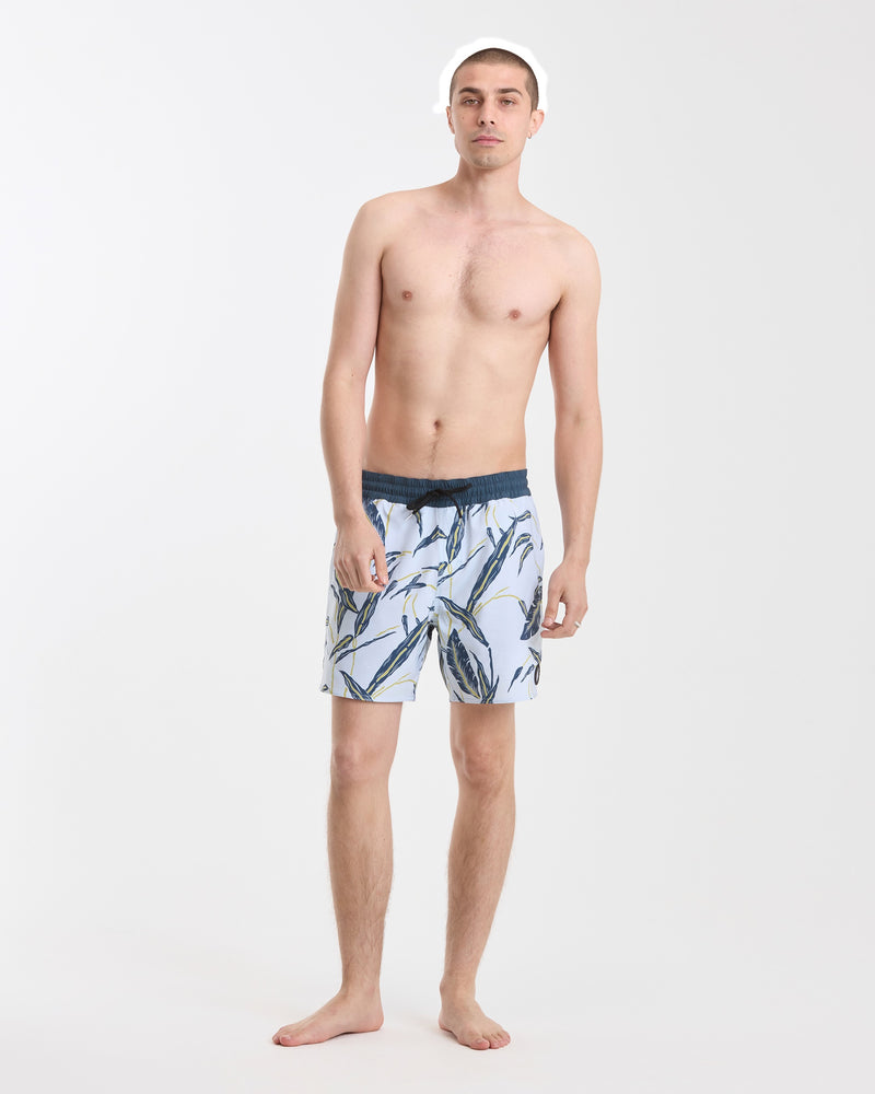 A young man stands barefoot against a plain white background, wearing Volcom Men's Lido Print 16" Trunks made from stretchy polyester-elastane with a back flap pocket, looking directly at the camera with a neutral expression.