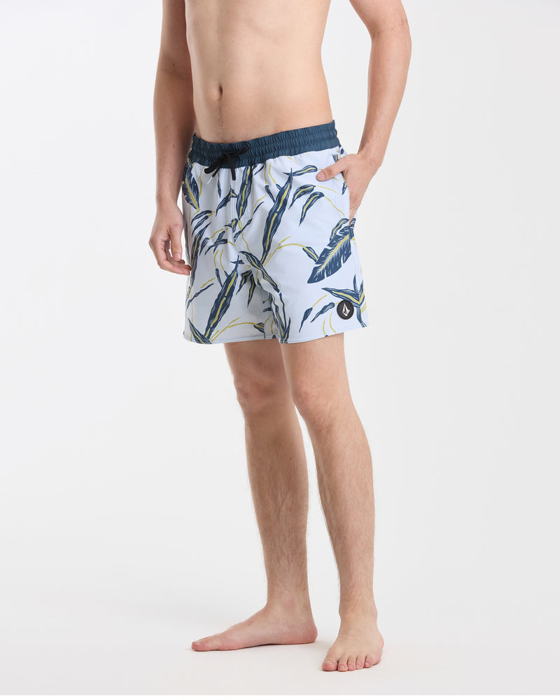 A man stands barefoot against a white background wearing Volcom’s Men's Lido Print 16" Trunks—light blue with a dark blue and yellow tropical leaf pattern, stretch polyester-elastane, dark blue waistband, back flap pocket, left hand in pocket.