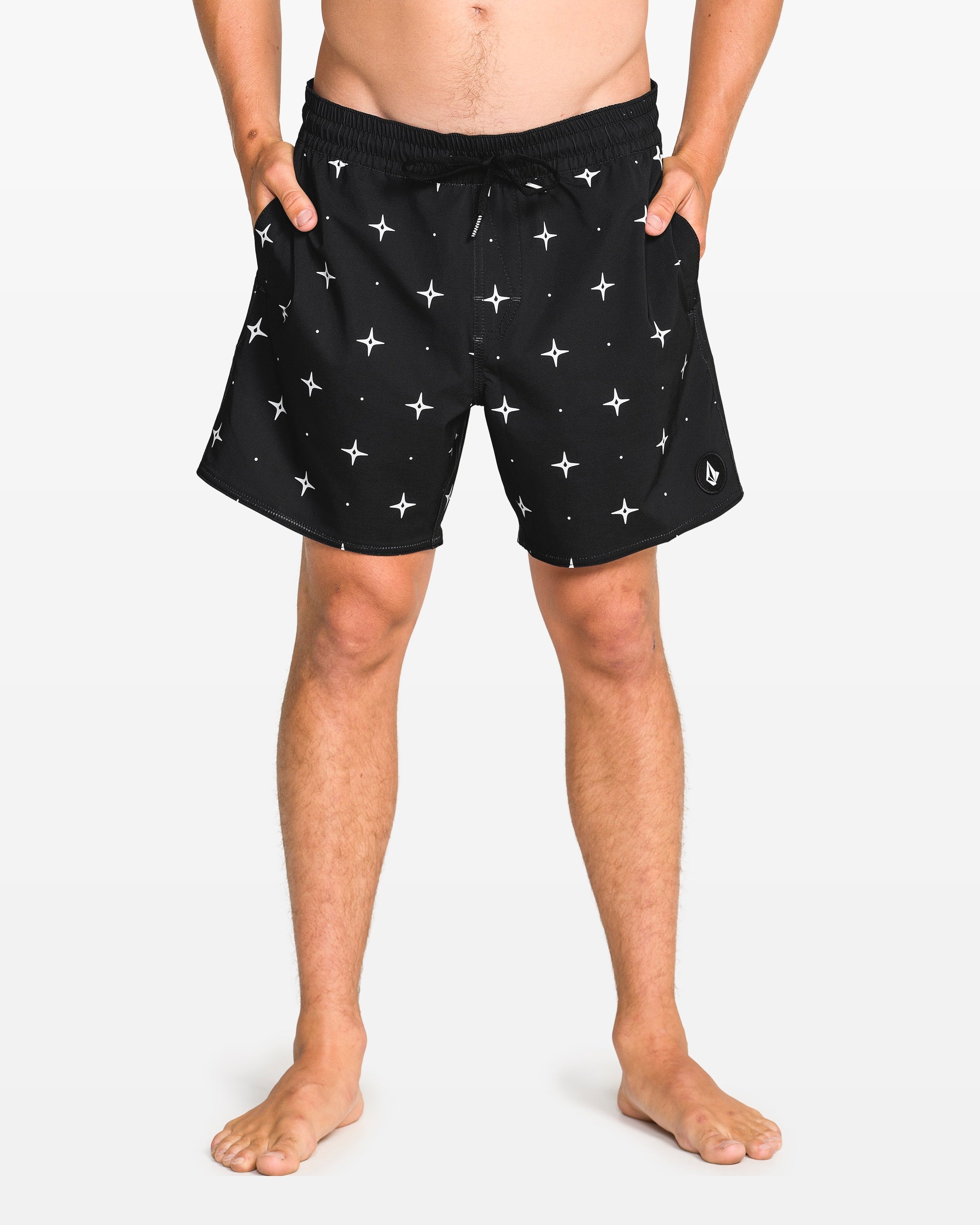 A person stands barefoot in Volcom Men's Lido Print 16" Trunk shorts with a white star print, elastic waistband with drawstring, 4-way stretch, and side pockets. The background is plain white.