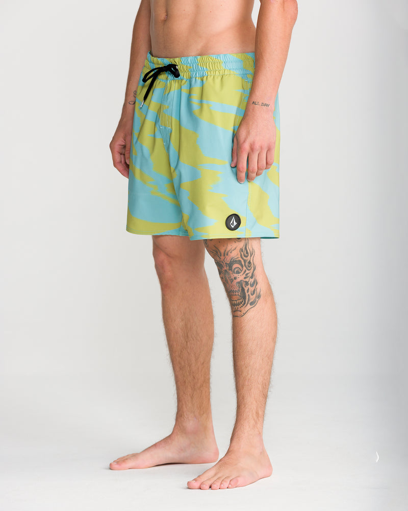 A person stands barefoot against a plain light background, showing a tattoo on their right thigh while wearing the Volcom Men’s Lido Print Trunk 16 swimwear in a blue and yellow camo pattern with a black drawstring and 4-way stretch fabric.