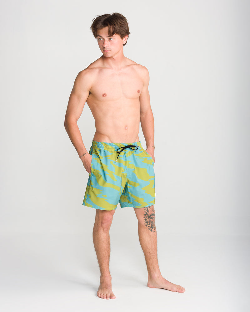 A young man stands barefoot against a plain background, wearing the Volcom Men’s Lido Print Trunk 16 swimwear in blue and green with a black drawstring. Featuring comfortable 4-way stretch, his hands are in his pockets as he looks slightly to the side.