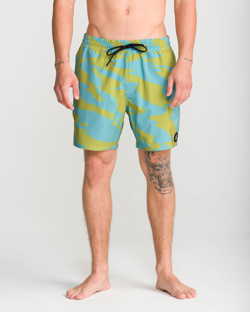 A person stands barefoot wearing Volcom Men’s Lido Print Trunk 16 swimwear in a green and blue pattern with a black drawstring. Their arms and part of their legs, including a visible tattoo, can be seen. Crafted from 4-way stretch fabric, these swim trunks provide comfort and flexibility. The background is plain white.