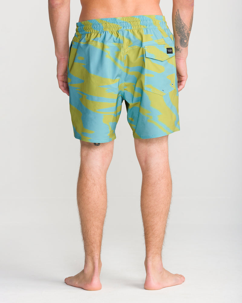 A person stands barefoot facing away, wearing Volcom’s Men’s Lido Print Trunk 16 swimwear in a blue and yellow pattern, featuring a back pocket. Only the lower back and legs are visible, highlighting the comfortable fit and 4-way stretch fabric. The background is plain and light-colored.
