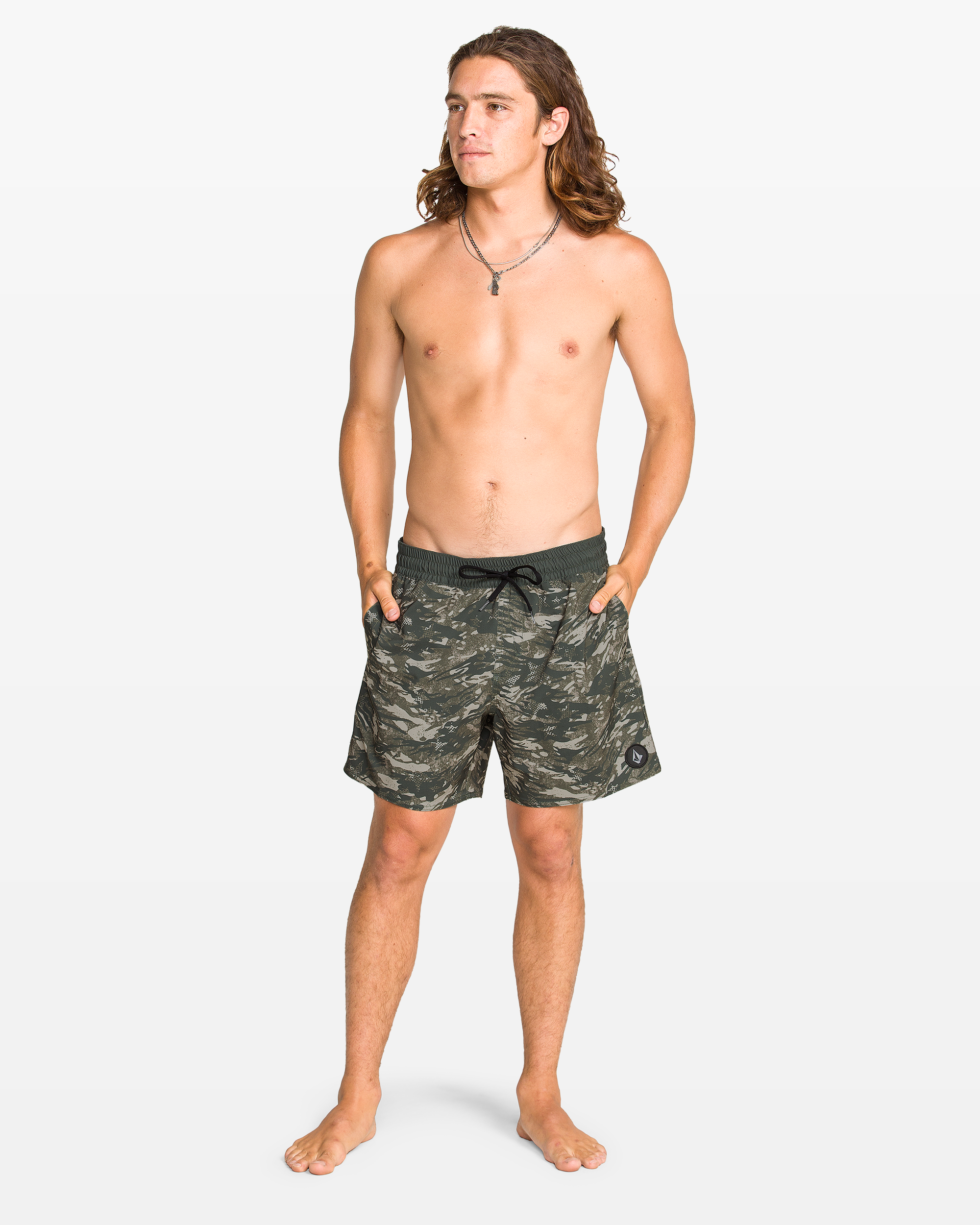 A barefoot, shirtless young man with long hair stands on a white background, wearing Volcom Men's Lido Print Trunks, featuring 4-way stretch camo fabric. He has his hands in his pockets and wears a necklace.