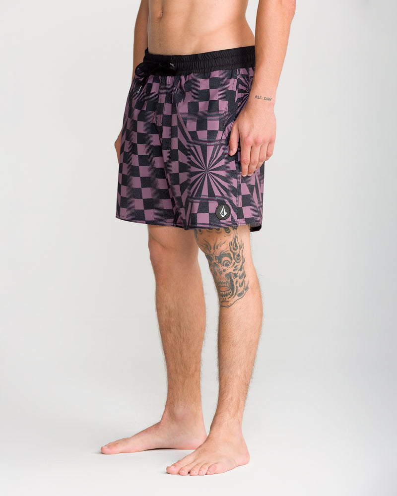 A man stands barefoot, wearing the Volcom Men’s Lido Print Trunk 16 in a striking black and purple checkered design with a geometric starburst pattern. Featuring 4-way stretch for enhanced comfort, the swim trunks allow ease of movement. A tattoo is visible on his left leg and part of his arm, while the simple, light-colored background draws attention to his look.