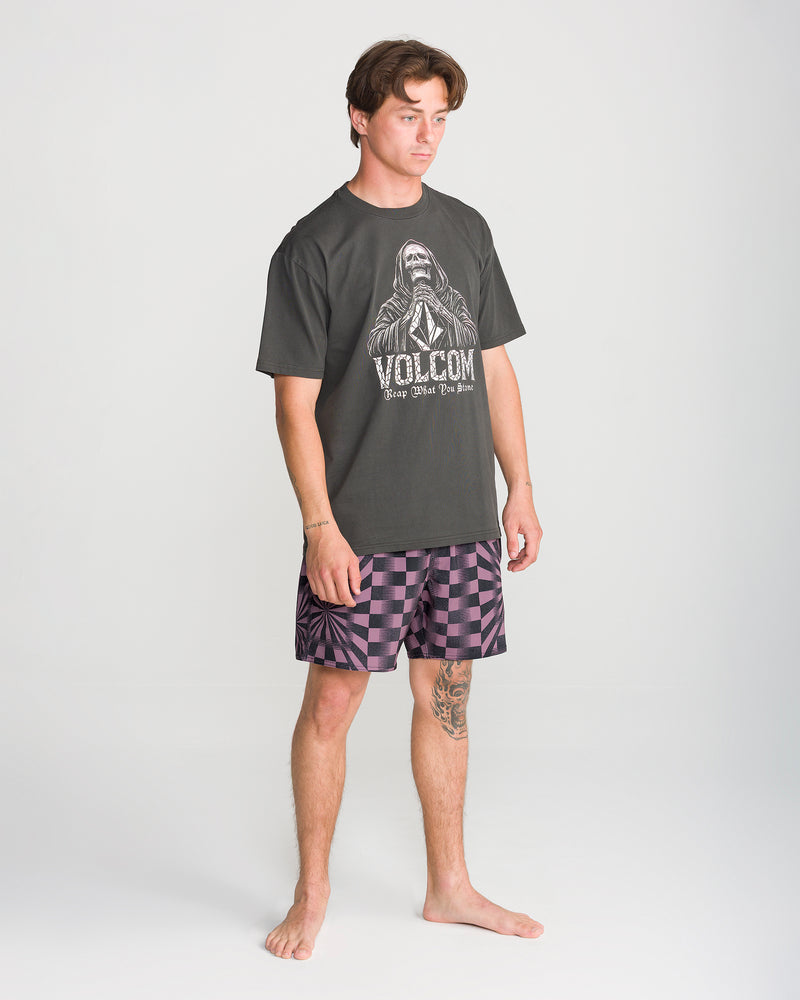A young man stands barefoot against a plain background, wearing a dark Volcom t-shirt and the Men’s Lido Print Trunk 16 shorts by Volcom, which feature a purple-black checkered print and 4-way stretch fabric. A tattoo is visible on his lower left leg.