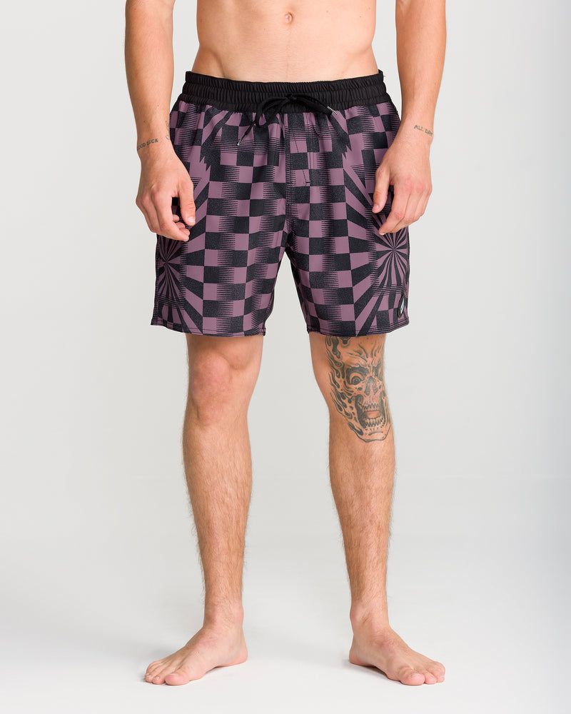 A person stands barefoot wearing the Volcom Men’s Lido Print Trunk 16 in a black and purple checkered pattern, featuring a Cinch Fly drawstring. Tattoos are visible on their right leg and arms, with a plain, light-colored background.