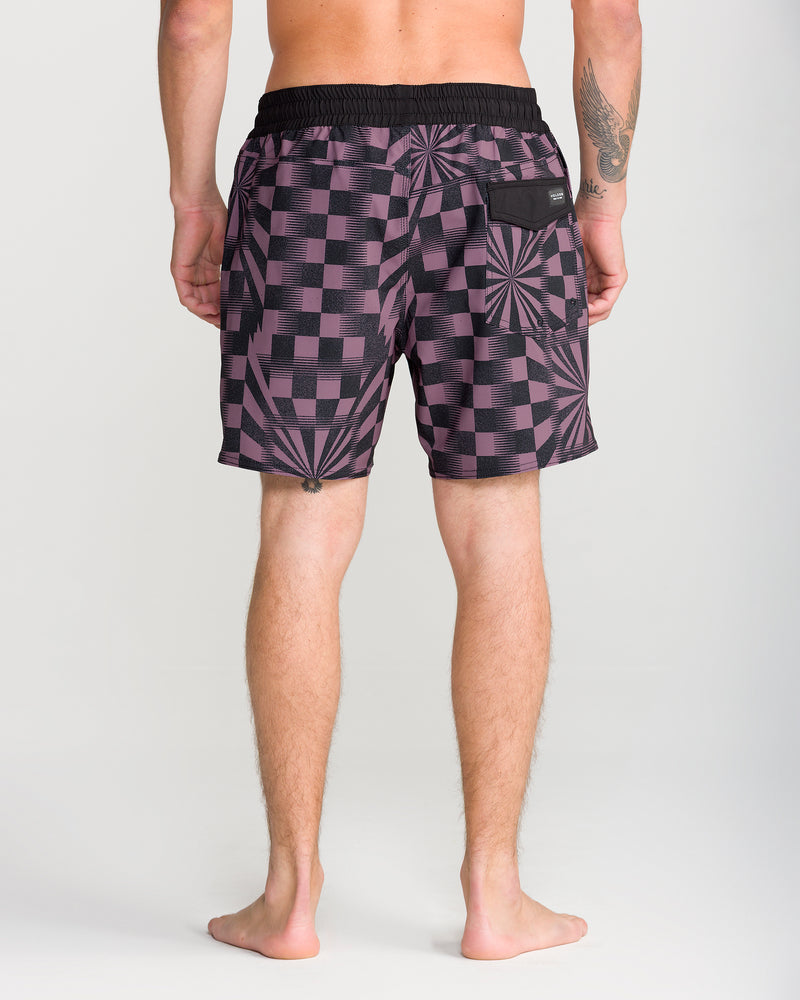A person stands barefoot, facing away, wearing the Volcom Men’s Lido Print Trunk 16 in a purple and black checkered design with geometric starburst patterns. The trunks feature a black waistband, a black pocket, and are made from comfortable 4-way stretch material. The person has a tattoo on their left arm.