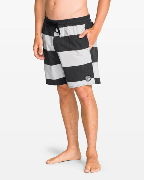 A person stands barefoot in Volcom Men's Psyche Cotton 17" Trunk—black and gray striped swimwear with a drawstring waist and one hand in the pocket, set against a plain white background.