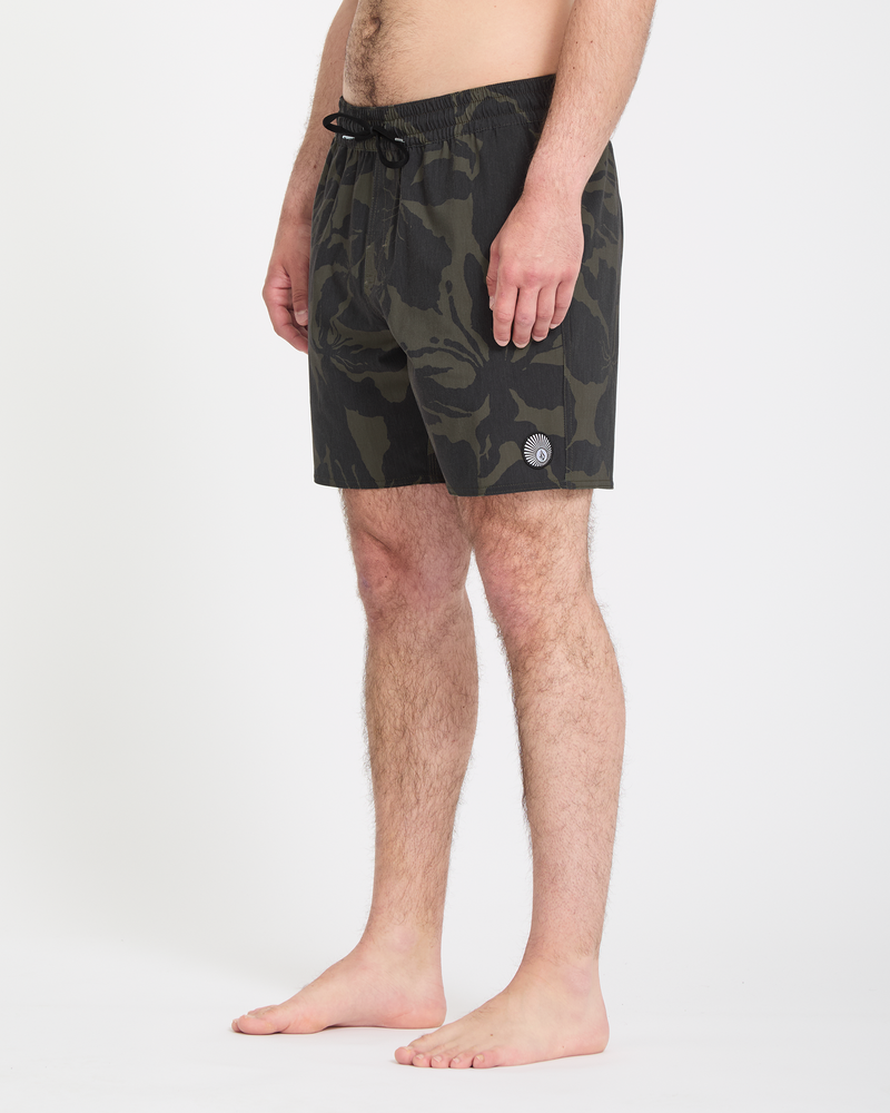 A man stands barefoot on a white background, wearing Volcom Men's Psyche Cotton Trunk 17 swim trunks with dark camouflage pattern, a black drawstring, and a small round patch near the hem. His torso is not visible.
