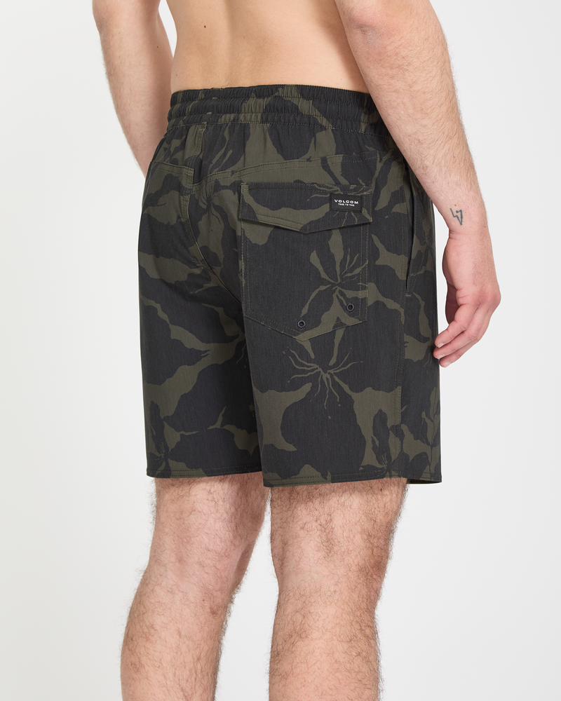 A man is seen from behind wearing Volcom Men's Psyche Cotton Trunk 17 shorts in black and dark green camo, featuring a back pocket; his upper body is bare with a small tattoo on his left arm.