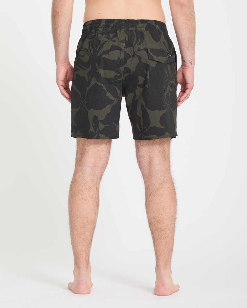 A barefoot person faces away from the camera, wearing Volcom Men's Psyche Cotton Trunk 17 shorts in a dark green and black camo pattern with an elastic waistband and back pocket. Their arms are relaxed at their sides.