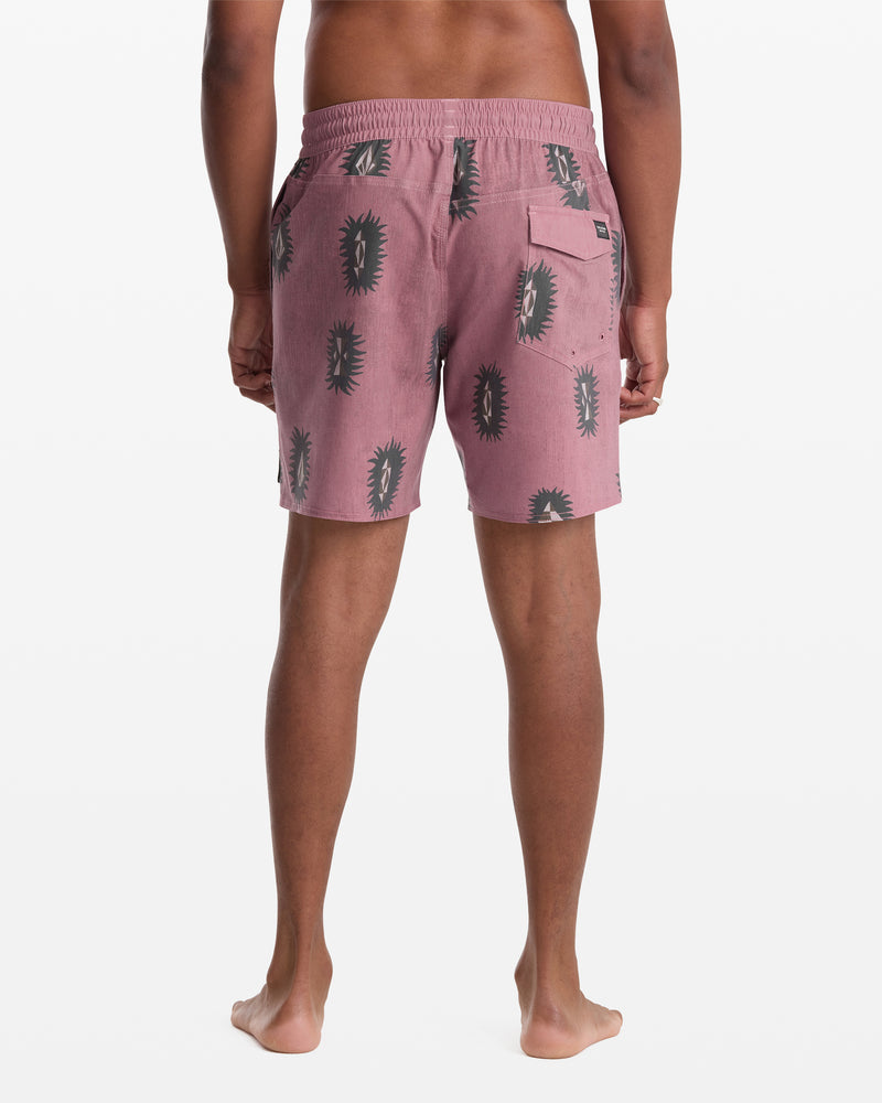 A person stands barefoot facing away, wearing Volcom Men's Psyche Cotton Trunk 17 shorts in pink with a black cactus pattern, featuring an elastic waistband and a back pocket.