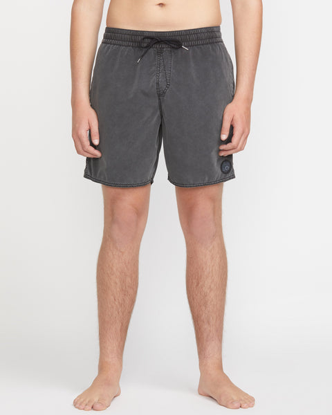 A person stands barefoot against a white background, wearing Volcom Men's Center Elastic Waist 17" Trunk swim shorts in dark gray, featuring recycled polyester, visible stitching, and a small circular patch near the left hem.