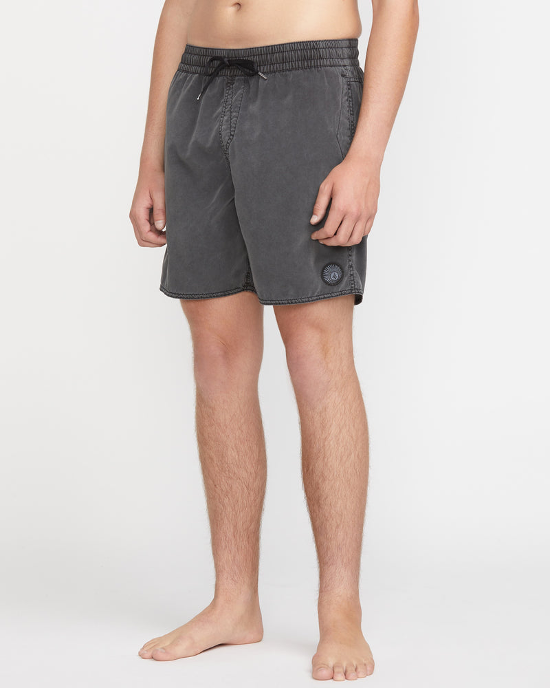 A person stands barefoot in Volcom's Men's Center Elastic Waist 17" Trunk, featuring a charcoal gray color, elastic waistband with drawstring, recycled polyester fabric with mechanical stretch, against a plain white background.