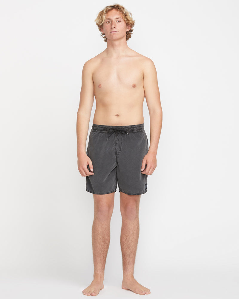 A young man with curly blond hair stands barefoot against a plain white background, wearing Volcom Men's Center Elastic Waist 17" Trunk in dark gray made from recycled polyester with mechanical stretch, facing forward neutrally with arms at his sides.