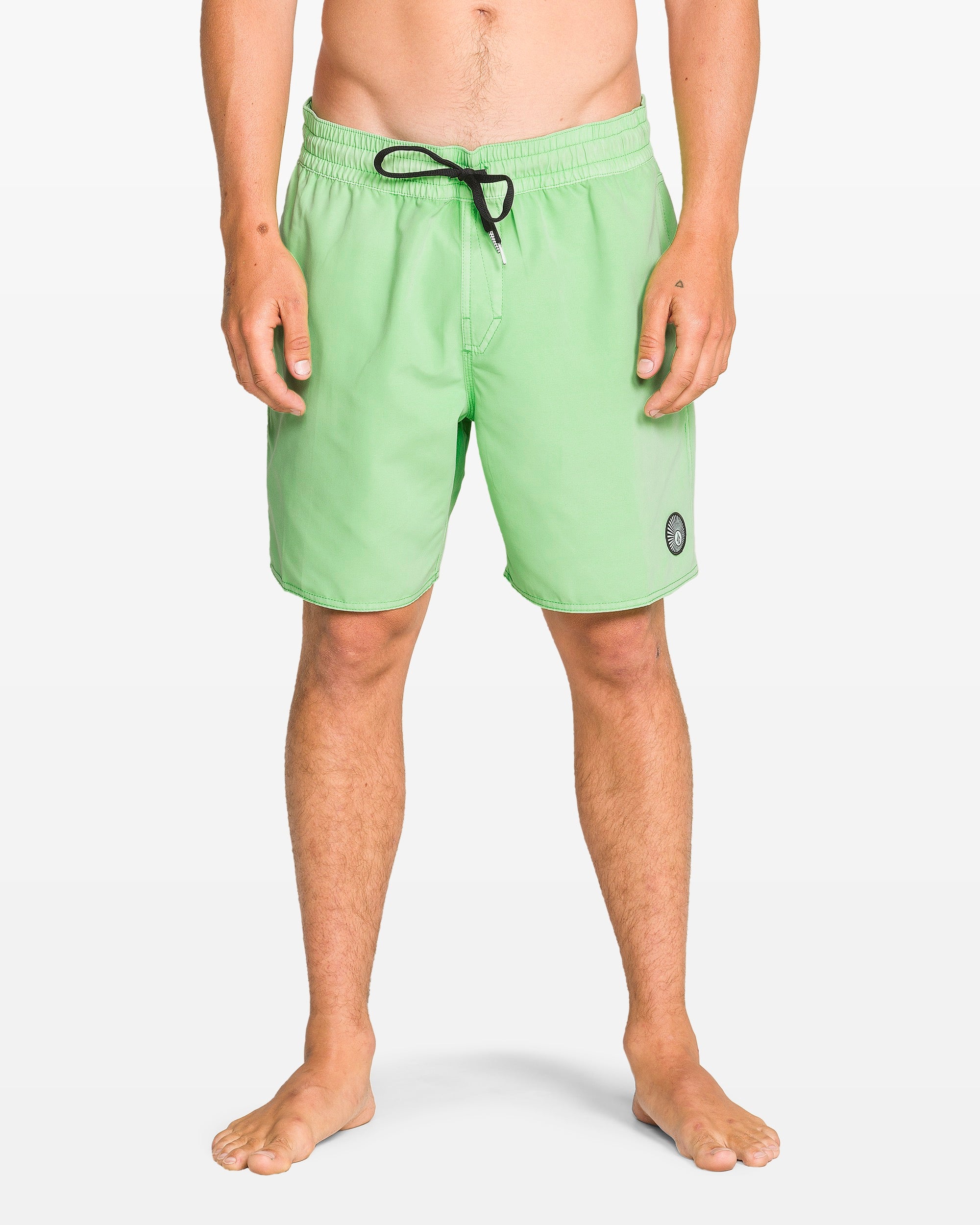 A barefoot person wears Volcom Men's Center Elastic Waist 17" Trunk swim shorts in light green, made from recycled polyester with 2-way stretch, featuring a black drawstring and small round logo on the left leg, against a plain white background.
