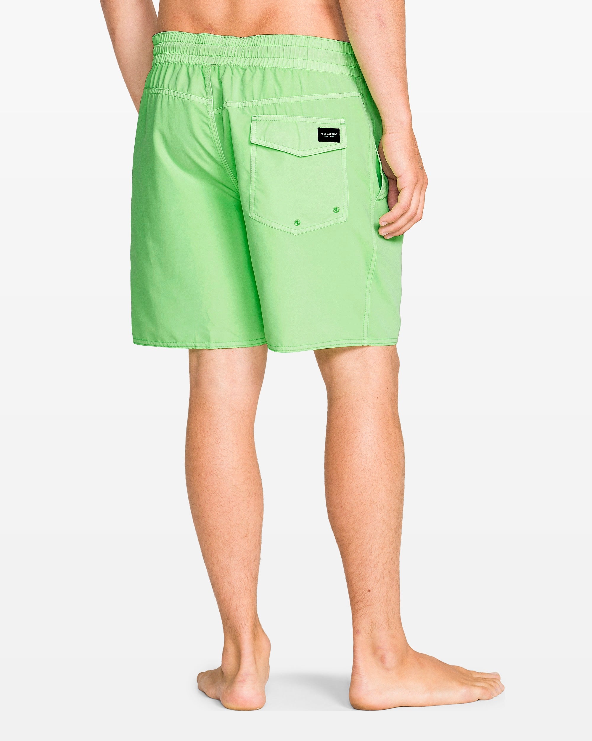 A barefoot person, shown waist down from the back, wears Volcom Men’s Center Elastic Waist 17" Trunk swim shorts in light green, made from recycled polyester with an elastic waistband and a flap back pocket.